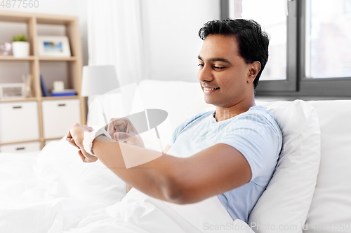 Image of happy indian man with smart watch in bed at home