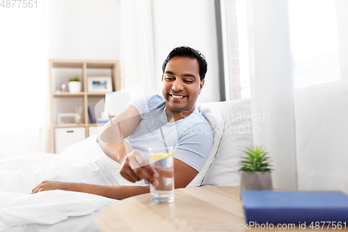 Image of happy indian man drinking water lying in bed