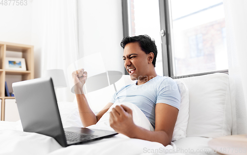 Image of happy indian man with laptop in bed at home