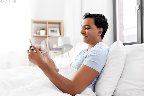 Image of happy indian man with smartphone in bed at home