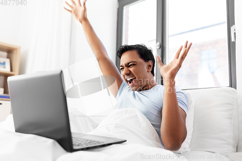 Image of happy indian man with laptop in bed at home