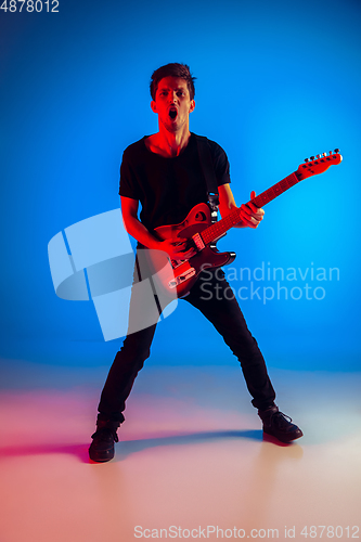 Image of Young caucasian musician playing guitar in neon light on blue background, inspired