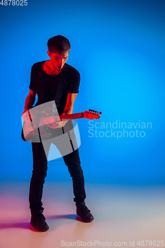 Image of Young caucasian musician playing guitar in neon light on blue background, inspired