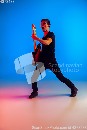 Image of Young caucasian musician playing guitar in neon light on blue background, inspired