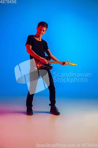 Image of Young caucasian musician playing guitar in neon light on blue background, inspired