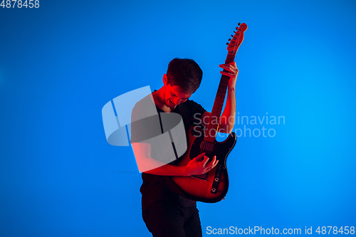 Image of Young caucasian musician playing guitar in neon light on blue background, inspired