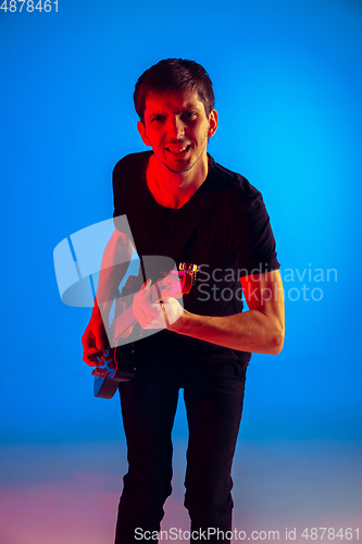 Image of Young caucasian musician playing guitar in neon light on blue background, inspired