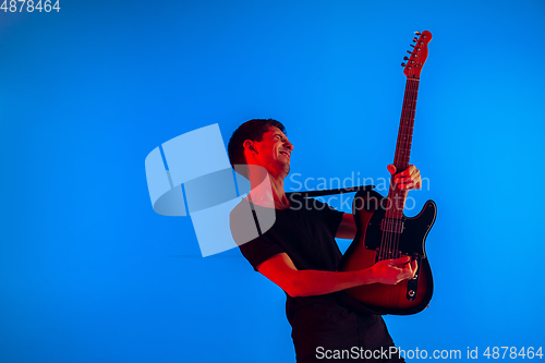 Image of Young caucasian musician playing guitar in neon light on blue background, inspired