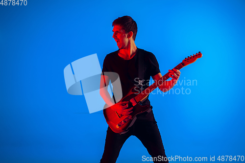 Image of Young caucasian musician playing guitar in neon light on blue background, inspired