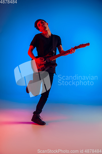 Image of Young caucasian musician playing guitar in neon light on blue background, inspired