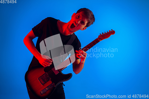 Image of Young caucasian musician playing guitar in neon light on blue background, inspired