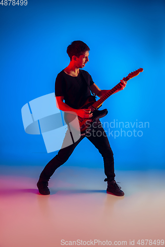 Image of Young caucasian musician playing guitar in neon light on blue background, inspired