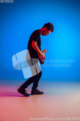 Image of Young caucasian musician playing guitar in neon light on blue background, inspired