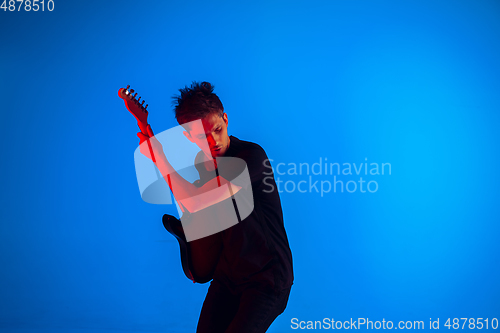 Image of Young caucasian musician playing guitar in neon light on blue background, inspired