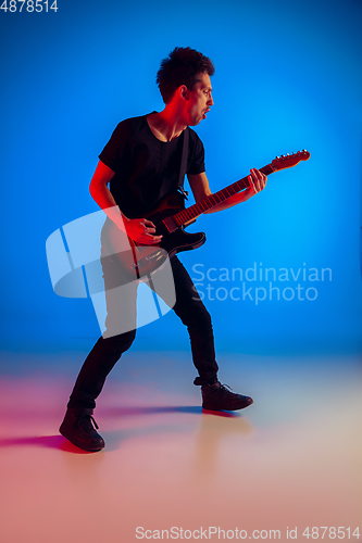 Image of Young caucasian musician playing guitar in neon light on blue background, inspired