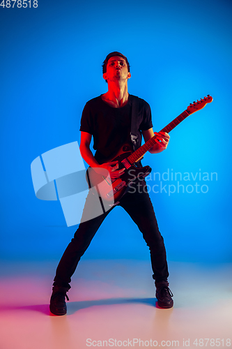 Image of Young caucasian musician playing guitar in neon light on blue background, inspired
