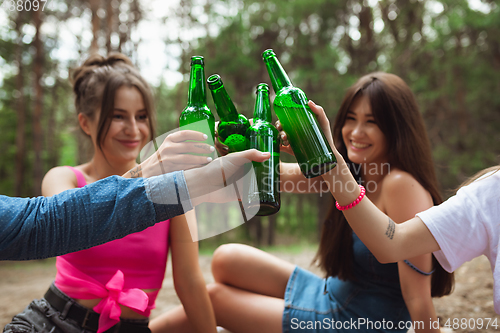 Image of Group of friends clinking beer bottles during picnic in summer forest. Lifestyle, friendship