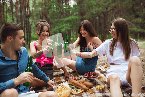 Image of Group of friends clinking beer bottles during picnic in summer forest. Lifestyle, friendship