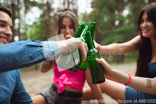 Image of Group of friends clinking beer bottles during picnic in summer forest. Lifestyle, friendship