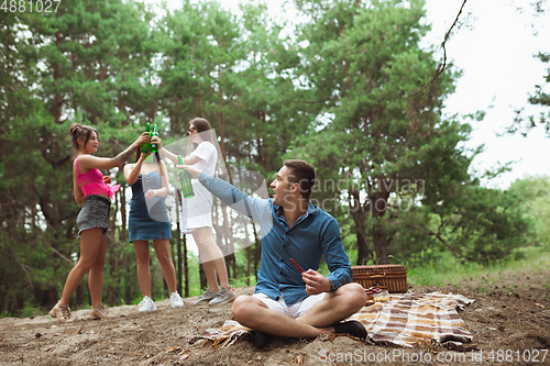 Image of Group of friends clinking beer bottles during picnic in summer forest. Lifestyle, friendship