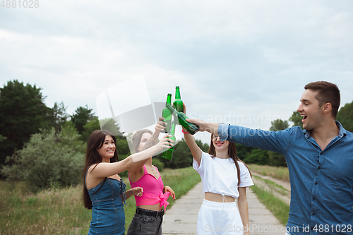 Image of Group of friends clinking beer bottles during picnic in summer forest. Lifestyle, friendship