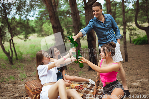 Image of Group of friends clinking beer bottles during picnic in summer forest. Lifestyle, friendship