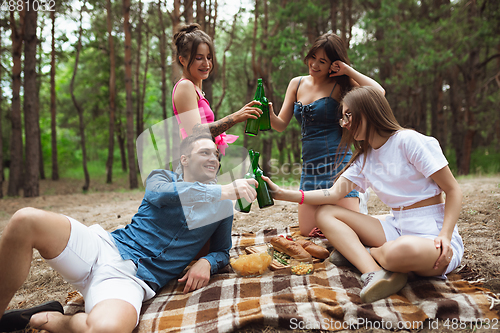 Image of Group of friends clinking beer bottles during picnic in summer forest. Lifestyle, friendship