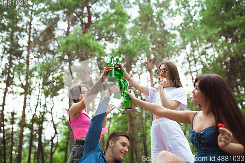 Image of Group of friends clinking beer bottles during picnic in summer forest. Lifestyle, friendship