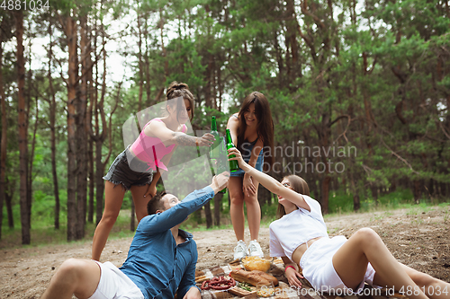 Image of Group of friends clinking beer bottles during picnic in summer forest. Lifestyle, friendship