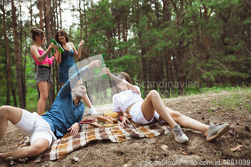 Image of Group of friends clinking beer bottles during picnic in summer forest. Lifestyle, friendship