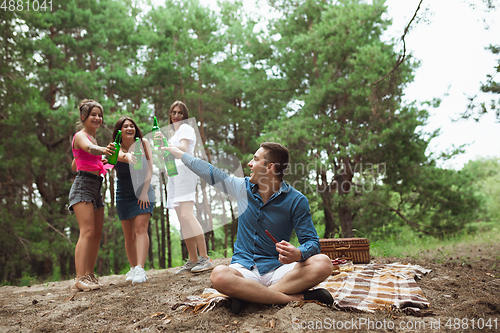 Image of Group of friends clinking beer bottles during picnic in summer forest. Lifestyle, friendship