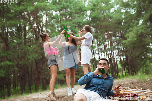 Image of Group of friends clinking beer bottles during picnic in summer forest. Lifestyle, friendship