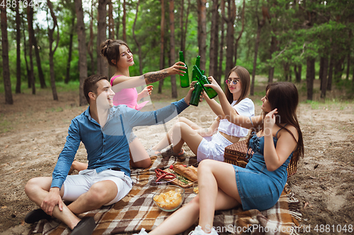 Image of Group of friends clinking beer bottles during picnic in summer forest. Lifestyle, friendship