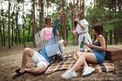 Image of Group of friends clinking beer bottles during picnic in summer forest. Lifestyle, friendship