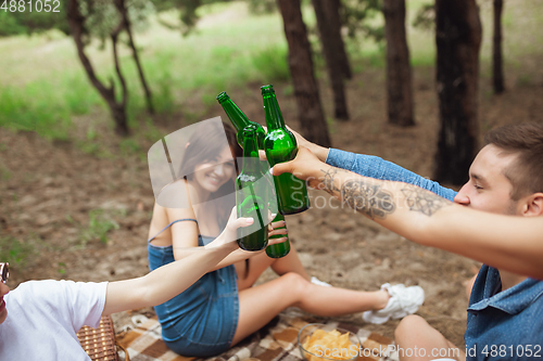 Image of Group of friends clinking beer bottles during picnic in summer forest. Lifestyle, friendship