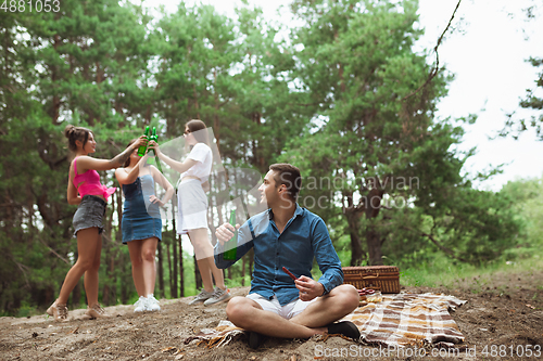 Image of Group of friends clinking beer bottles during picnic in summer forest. Lifestyle, friendship