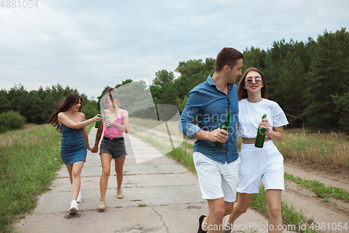 Image of Group of friends clinking beer bottles during picnic in summer forest. Lifestyle, friendship