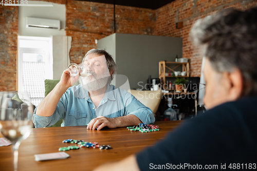 Image of Two happy mature friends playing cards and drinking wine