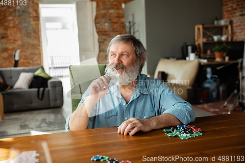 Image of Senior man playing cards and drinking wine with friends, looks happy