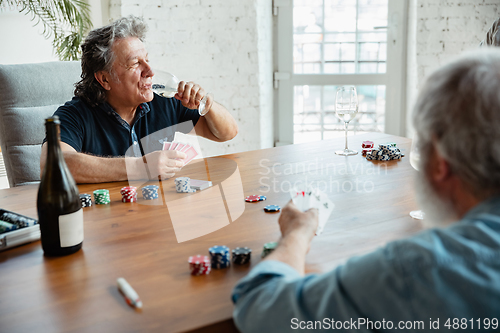 Image of Two happy mature friends playing cards and drinking wine