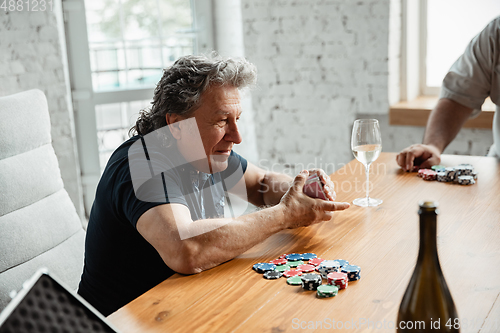 Image of Senior man playing cards and drinking wine with friends, looks happy