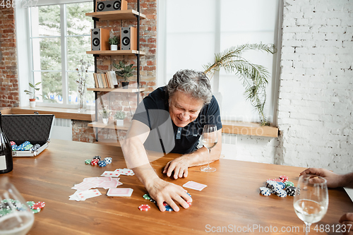 Image of Senior man playing cards and drinking wine with friends, looks happy