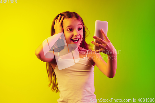 Image of Little caucasian girl\'s portrait isolated on gradient background in neon light. Concept of human emotions, facial expression, modern gadgets and technologies. Copyspace.