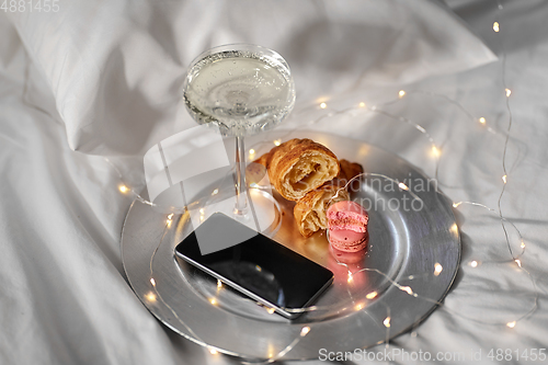 Image of champagne glass, croissants and smartphone in bed