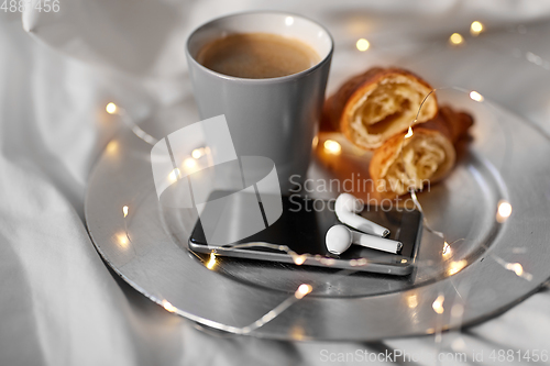 Image of smartphone, earphones, coffee and croissant in bed