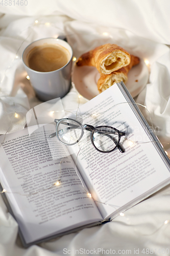 Image of croissants, cup of coffee, book and glasses in bed