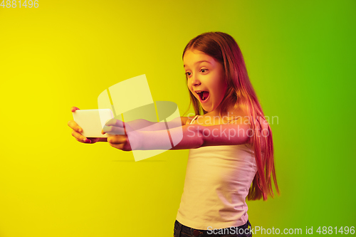 Image of Little caucasian girl\'s portrait isolated on gradient background in neon light. Concept of human emotions, facial expression, modern gadgets and technologies. Copyspace.