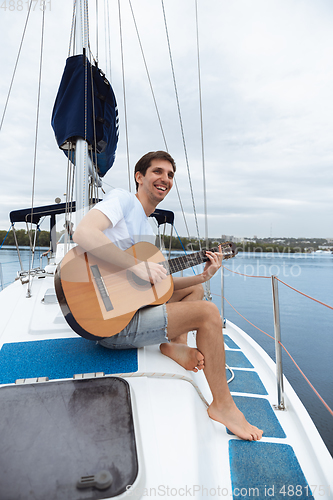 Image of Young cheerful man playing guitar at boat party outdoor, smiling and happy