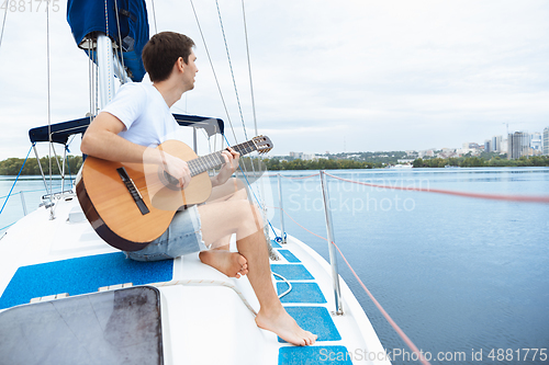 Image of Young cheerful man playing guitar at boat party outdoor, smiling and happy