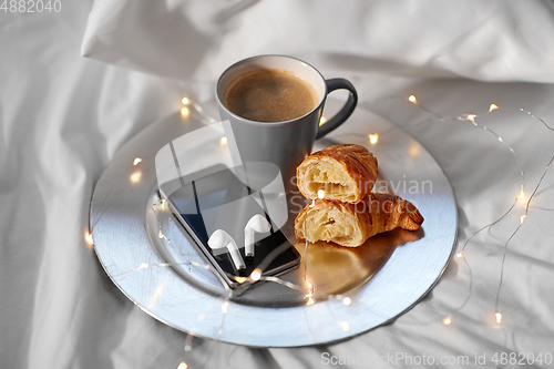 Image of smartphone, earphones, coffee and croissant in bed